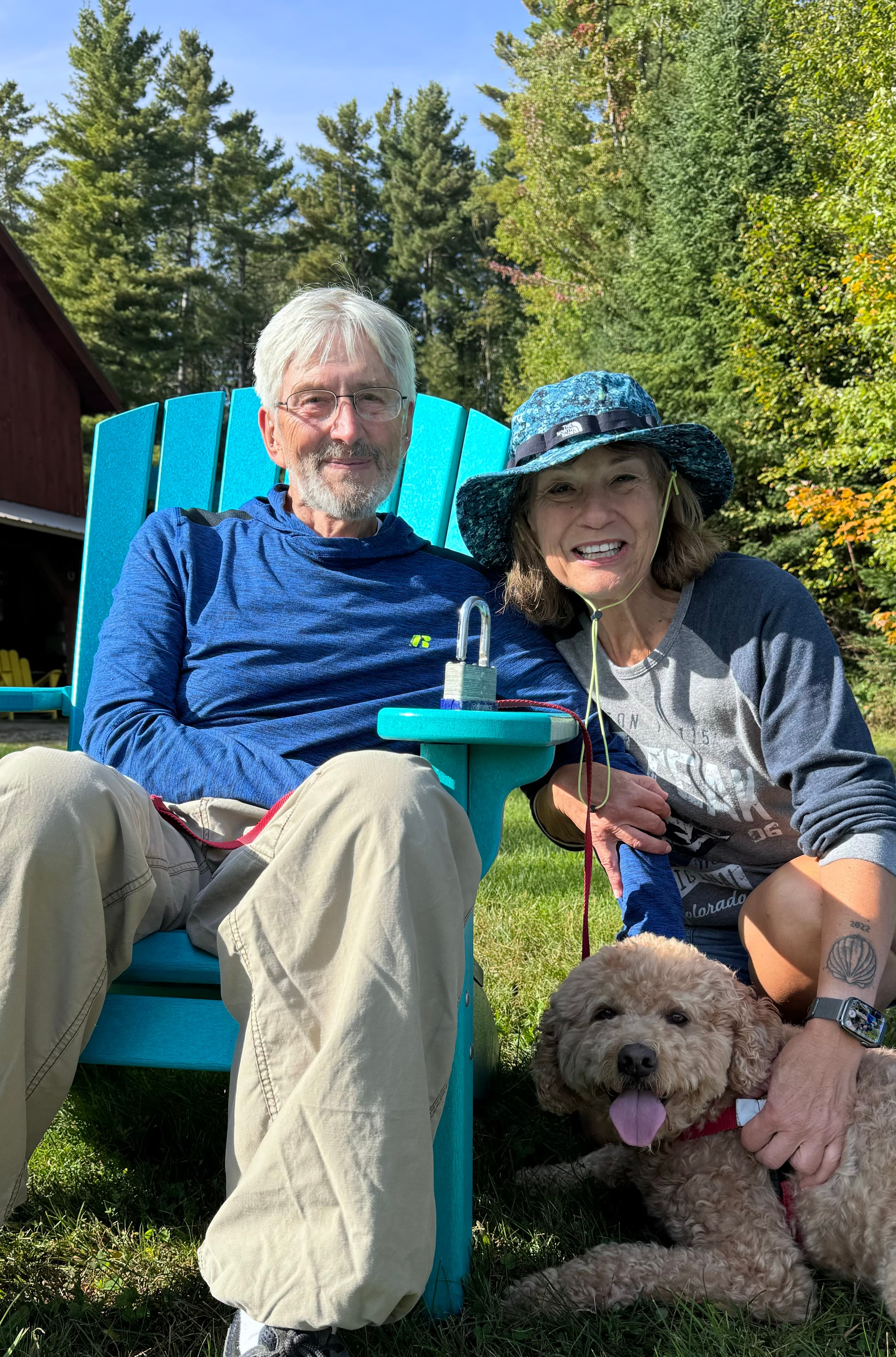 A man and a woman smile while sitting on bright blue chairs with a dog at their feet in a forested setting.