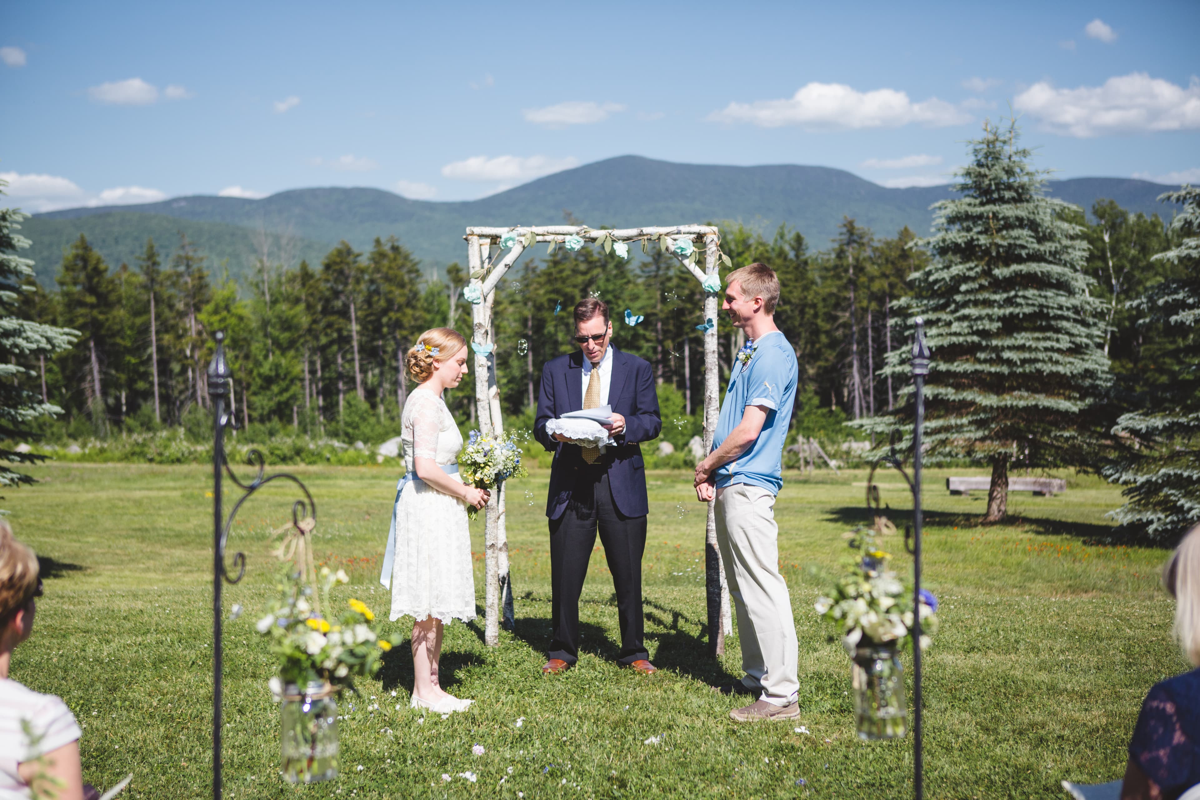 A couple stands under a floral arch while exchanging vows during their outdoor wedding ceremony, with mountains in the background.