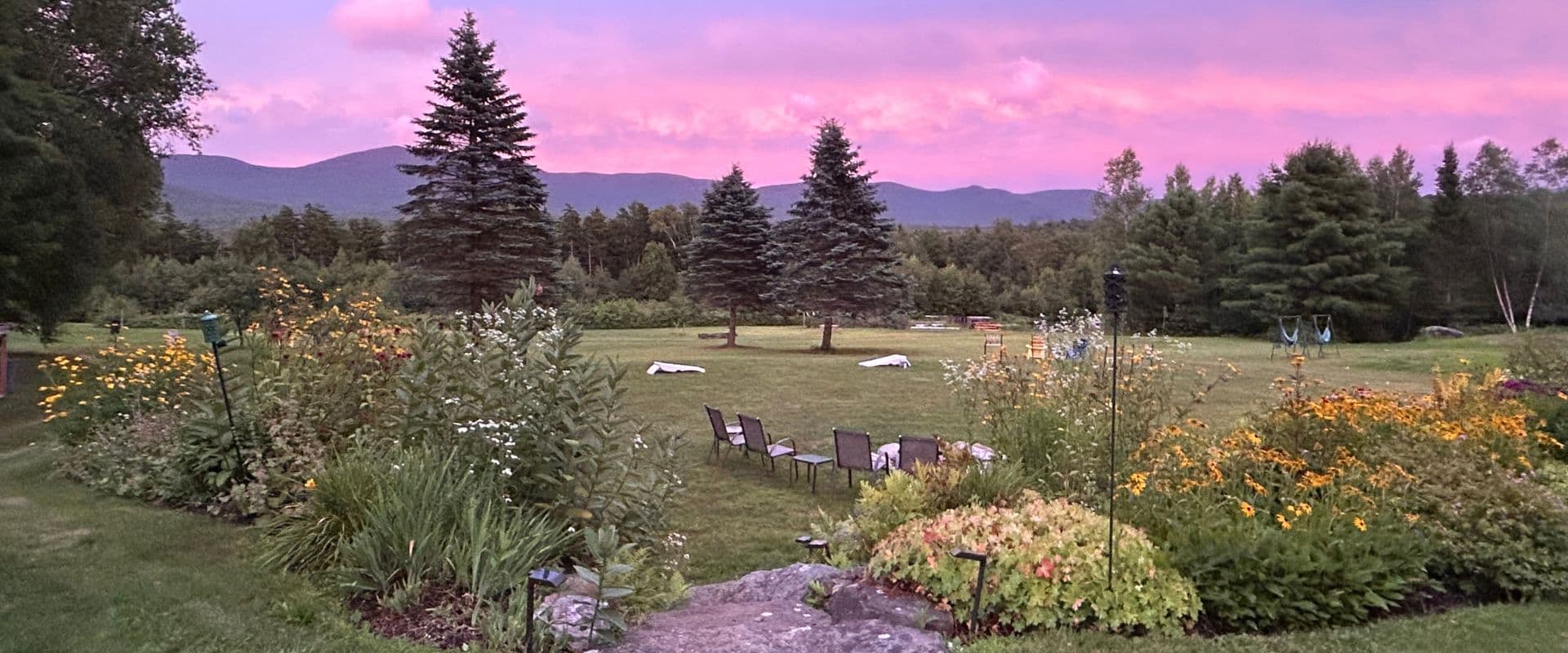 A lush garden with colorful flowers and lounge chairs overlooks rolling hills under a pink-hued sky.