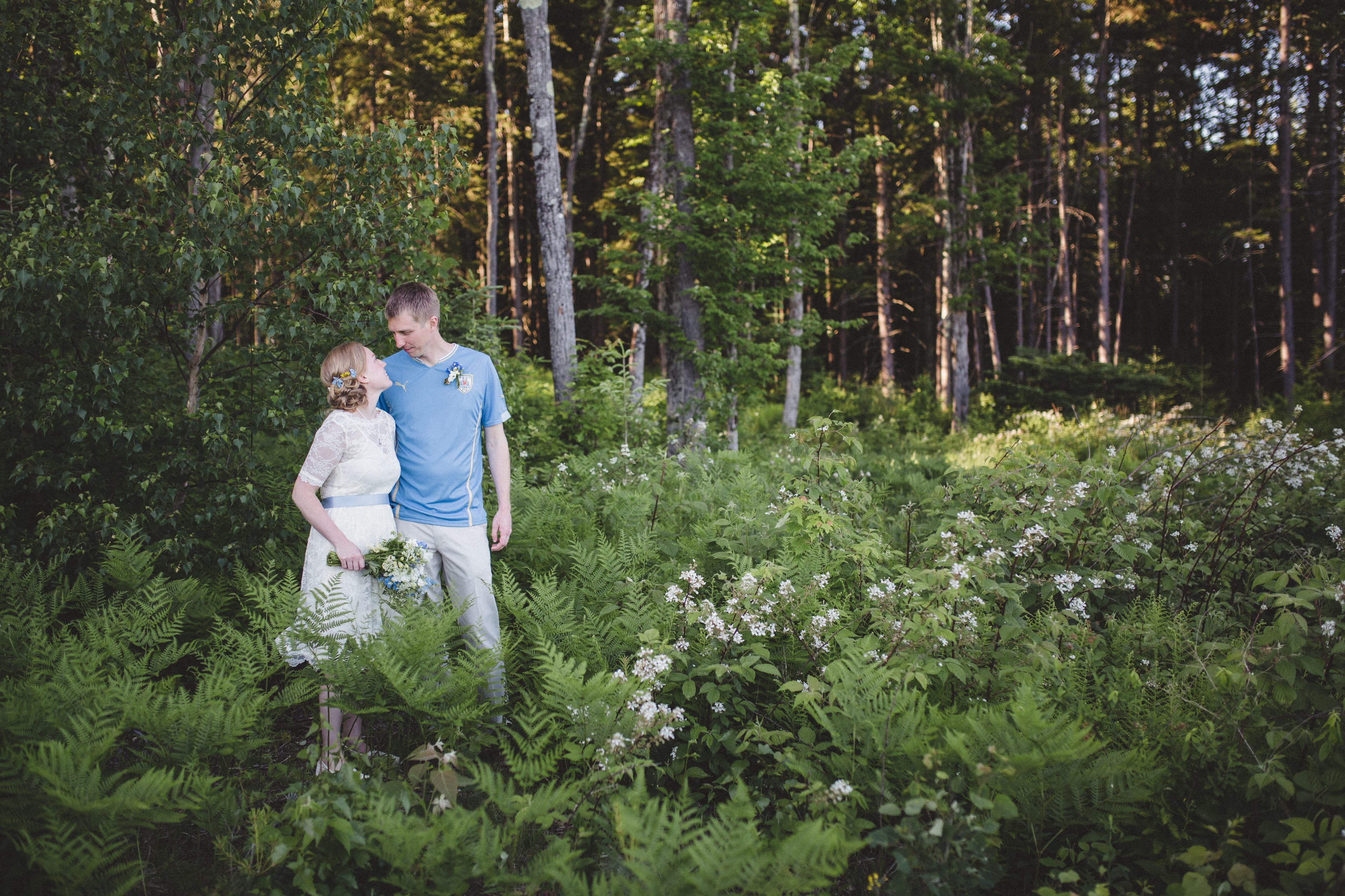 A couple stands affectionately among lush green ferns and wildflowers in a forest setting.