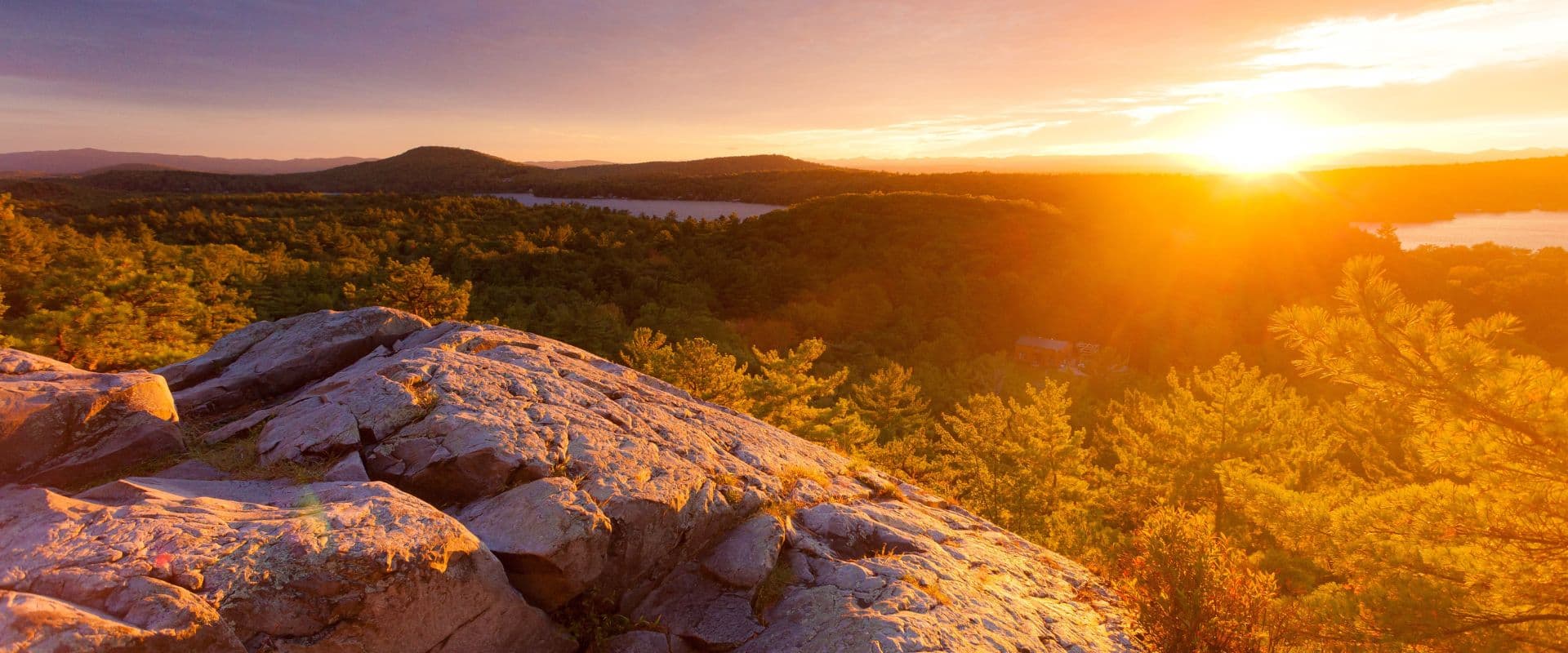 Sunrise over a picturesque landscape of mountains and forests, with rocky foreground.