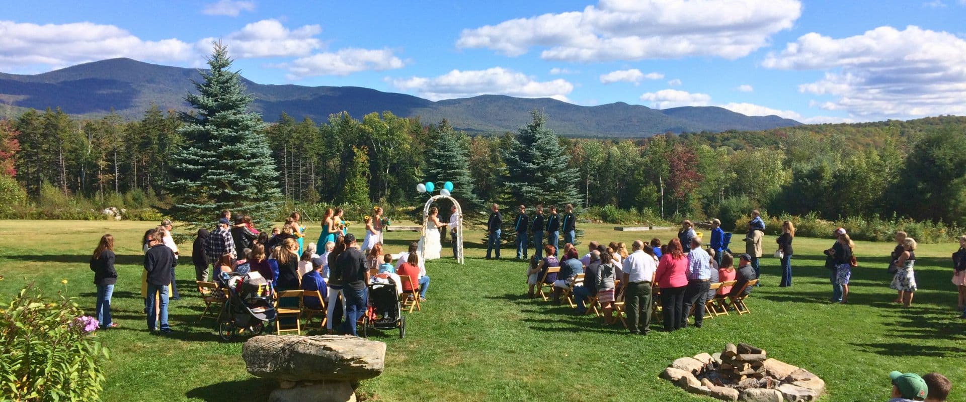 A wedding ceremony is taking place outdoors with guests seated on grass surrounded by trees and mountains.