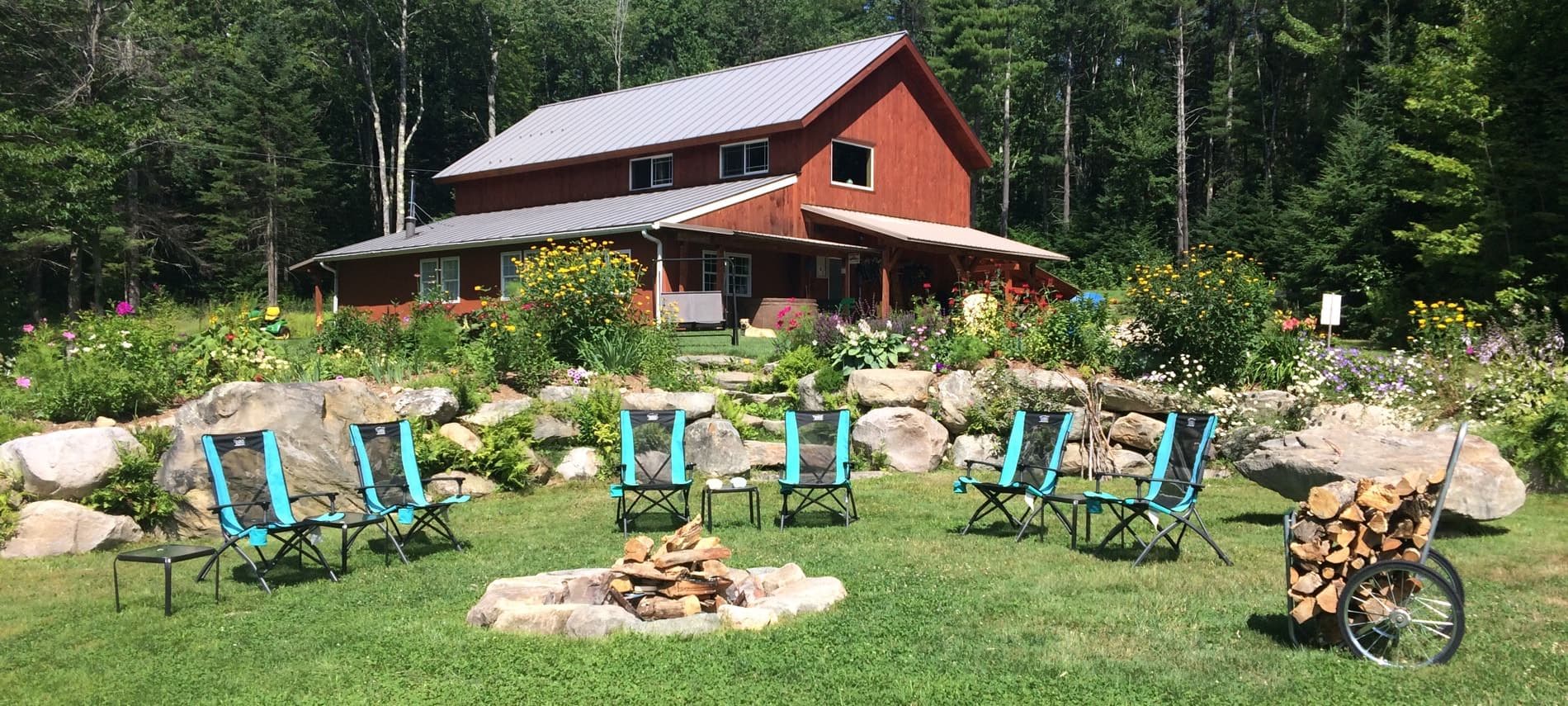 A red house is surrounded by a garden, with chairs arranged around a stone fire pit.