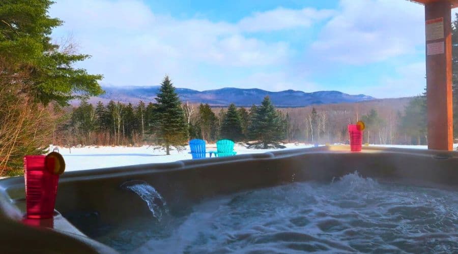 A hot tub with bubbles overlooks a snowy landscape and distant mountains, with colorful chairs and drinks nearby.