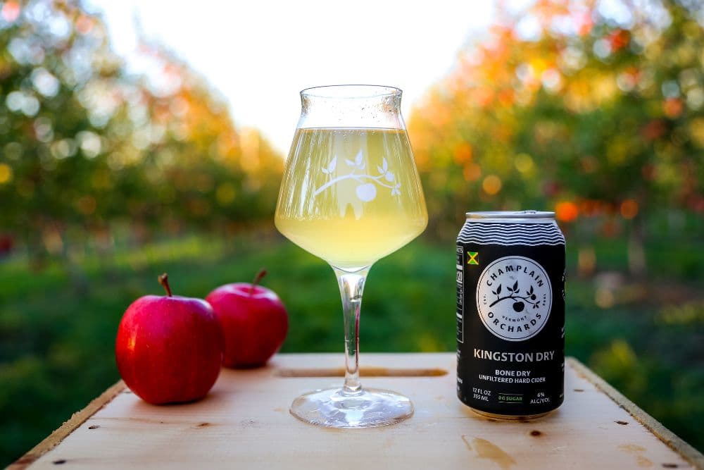 A glass of Kingston Dry cider sits next to a can and two red apples, with an orchard backdrop.