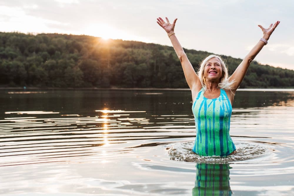 A joyful woman in a colorful swimsuit stands in a lake, raising her arms in celebration as the sun sets in the background.