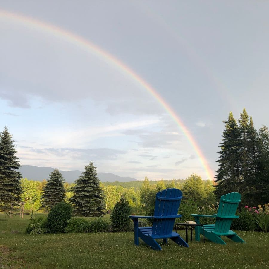 Two Adirondack chairs in a lush garden under a rainbow arching across a cloudy sky.
