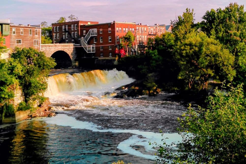 A waterfall cascades into a river beside brick buildings and greenery.