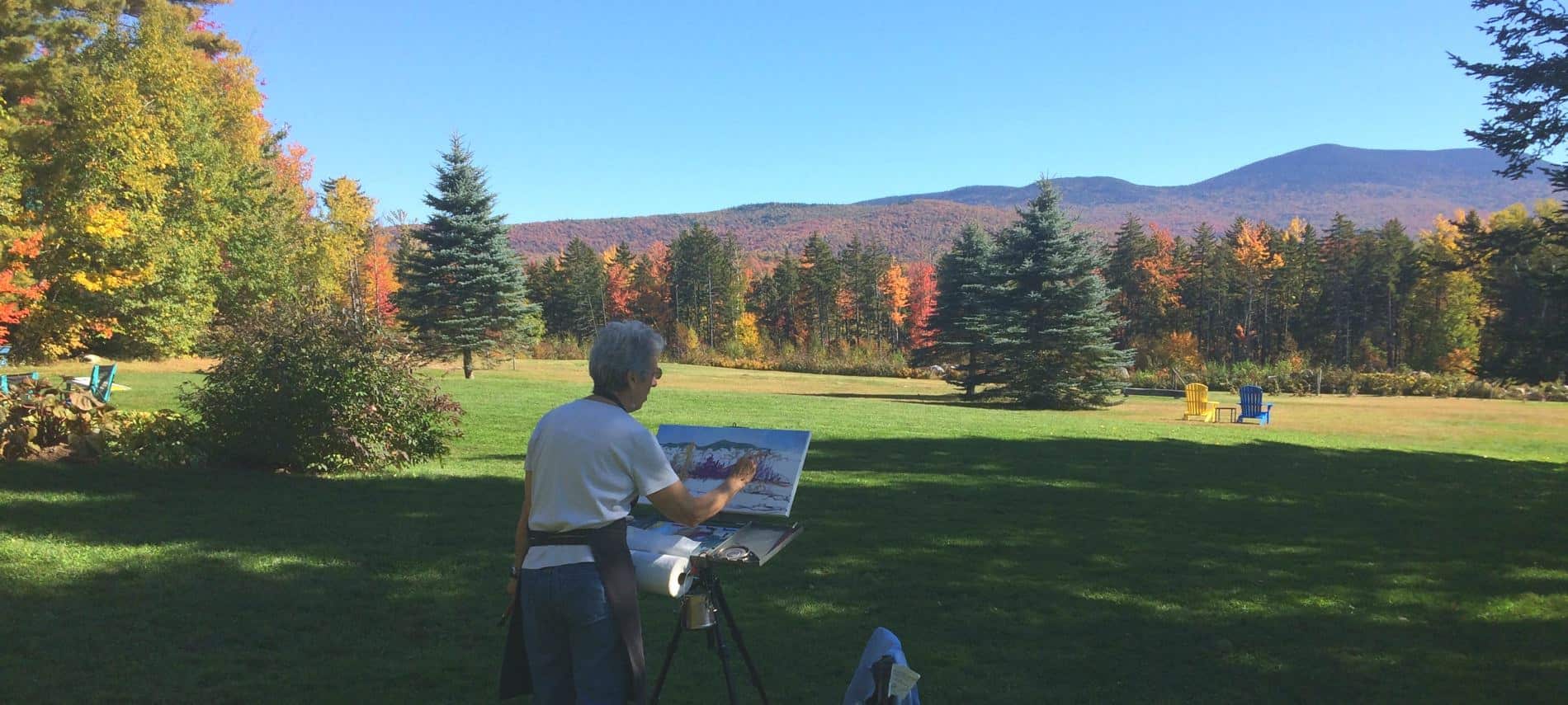 Person standing outside in the grass painting the view of green grass, colorful trees in the fall and distant hills